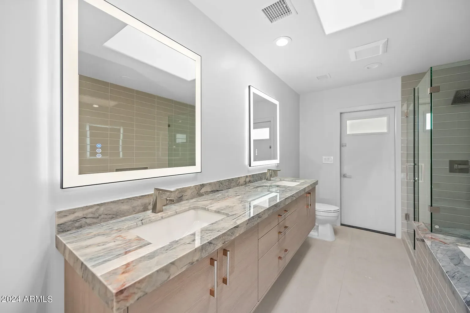 Marble Double Vanity: Book-matched slab vanity with backlit mirrors and a frameless glass enclosure beyond.